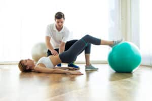 Physiotherapist helping patient to do exercise on fitness ball in physio room.